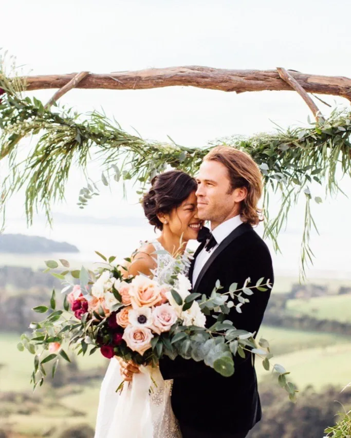 Bride and groom embracing under a driftwood and floral arch with the Hauraki Gulf behind